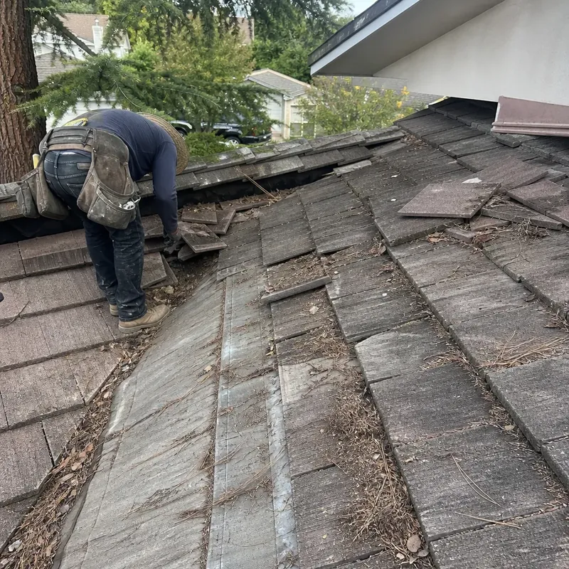 Worker inspecting an exposed tile roof valley with displaced tiles and accumulated debris.