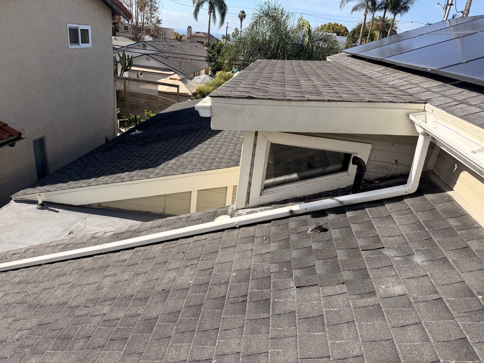 Dana Point roof overview with shingle slopes, dormer window, downspout, existing solar panels and a coastal view beyond the palms.