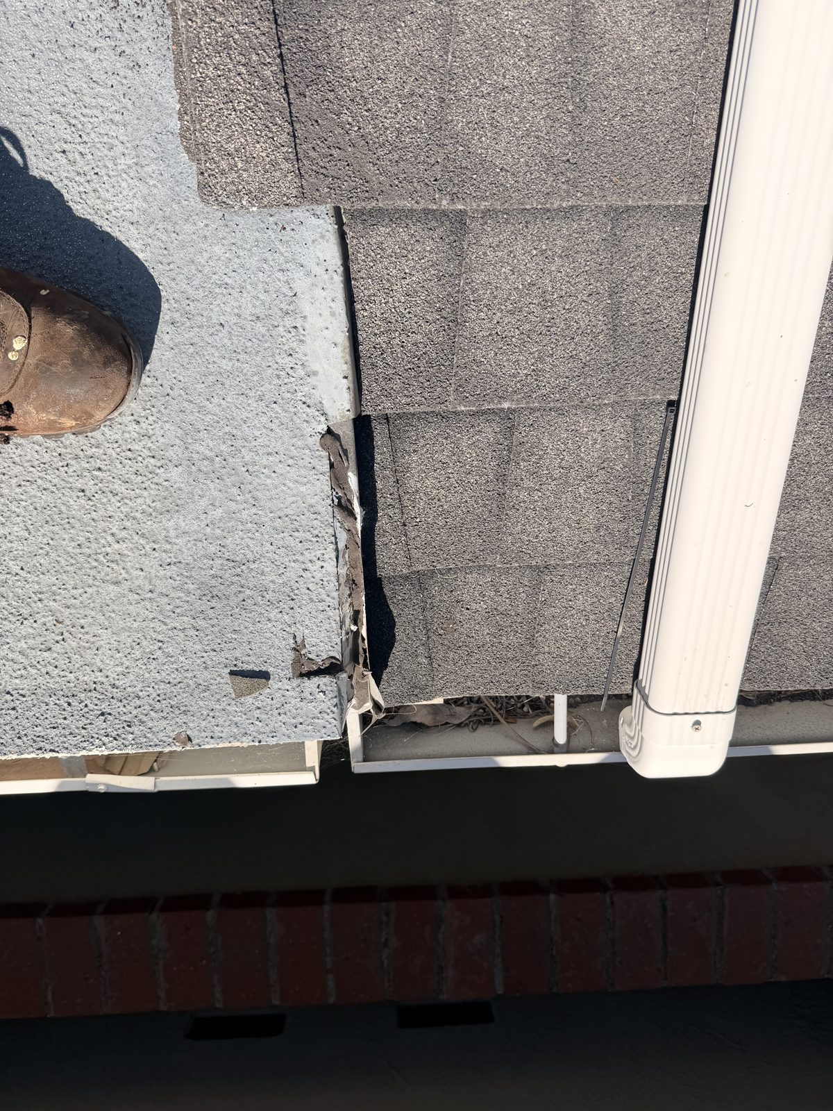 Damaged edge where the flat-roof membrane meets asphalt shingles beside the gutter and downspout.